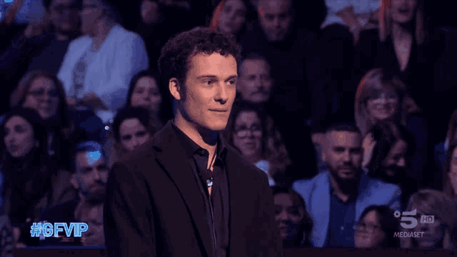 a man in a black suit stands in front of a crowd with the hashtag #gfvip on the screen