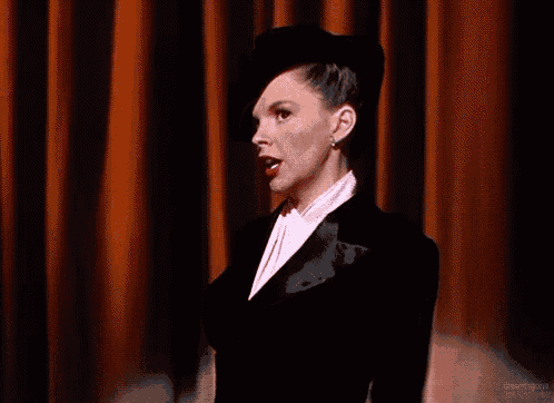 a woman wearing a tuxedo and a hat is waving her hand in front of an orange curtain .
