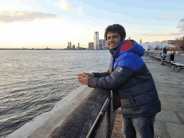 a man wearing a jacket that says ' columbia ' on it leans against a railing overlooking a body of water