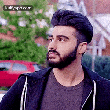 a man with a beard and a black jacket is standing in front of a red car .