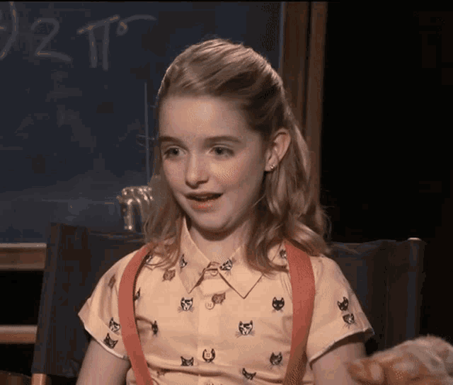a young girl wearing a shirt with cats on it sits in front of a blackboard that says 1/2 tp