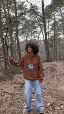 a woman wearing a brown hoodie and jeans is standing in the woods