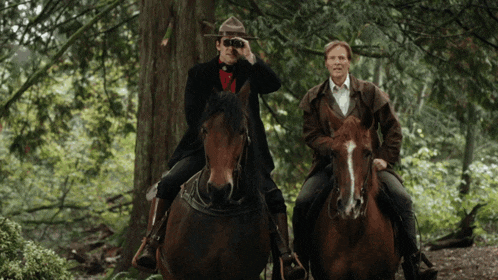 two men are riding horses through the woods and one is looking through binoculars