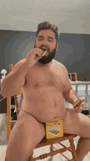 a man without a shirt is sitting on a chair eating a snickers bar