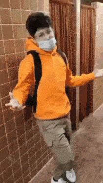 a person wearing a mask and an orange hoodie stands in a bathroom