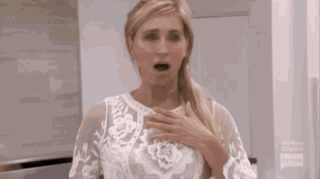 a woman in a white lace dress is holding her chest and making a surprised face .