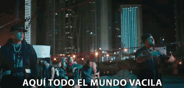 a man in a baseball cap is standing in front of a city at night with the words aqui todo el mundo vacila below him