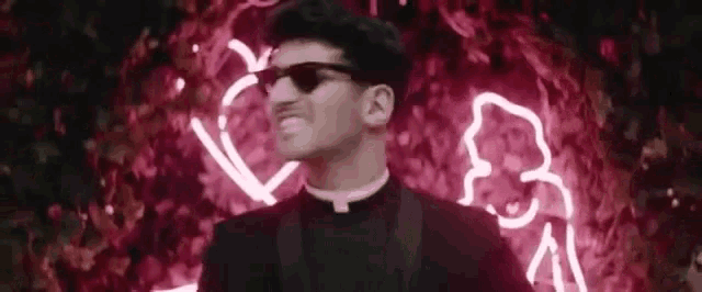a priest wearing sunglasses and a tuxedo is smiling in front of a neon sign .