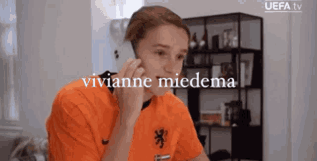a woman in an orange shirt is talking on a cell phone and the name vivianne miedema is on the screen