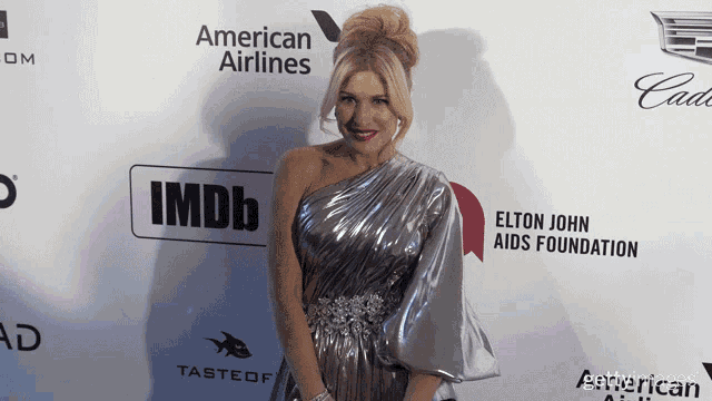 a woman in a silver dress stands on a red carpet sponsored by american airlines