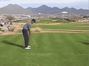 a man is swinging a golf club at a golf ball on a golf course with mountains in the background .