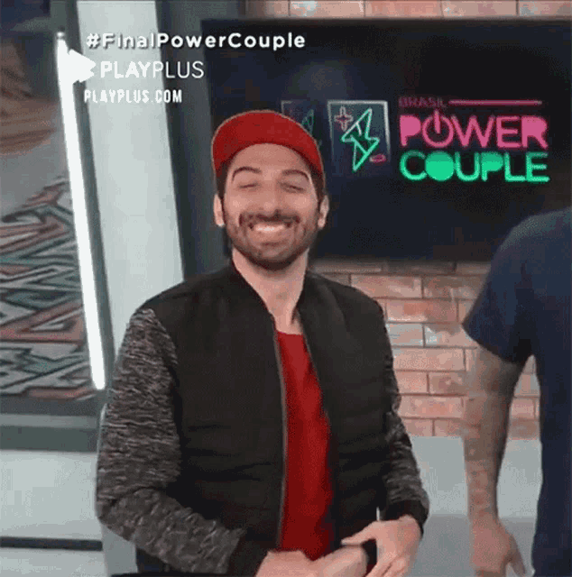 a man wearing a red hat and a black jacket is smiling in front of a screen that says power couple