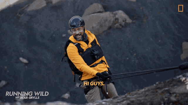 a man in a yellow jacket says hi guys as he climbs a mountain