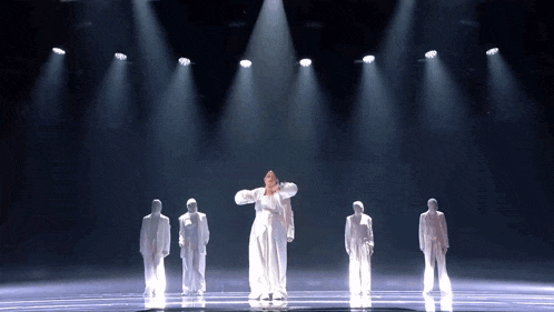a woman in a white dress is dancing on a stage with a group of people in white suits