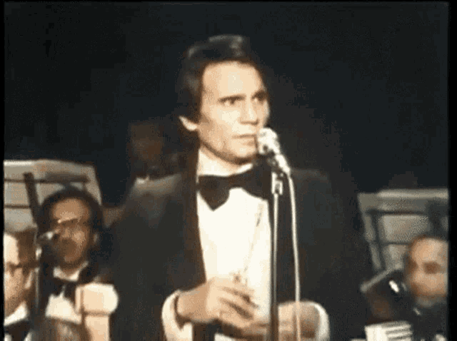 a man in a tuxedo sings into a microphone in front of an orchestra