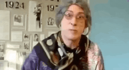 an elderly woman wearing a scarf and glasses is standing in front of a wall with pictures on it .