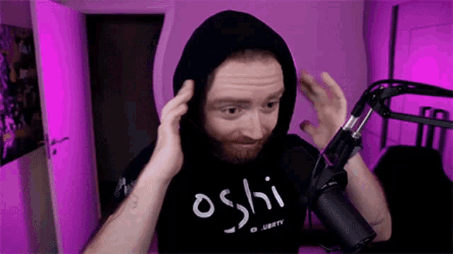 a man with a beard wearing a hoodie is standing in front of a microphone in a room .