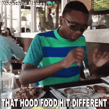 a young boy is sitting at a table with a plate of food and a caption that says that hood food hit different