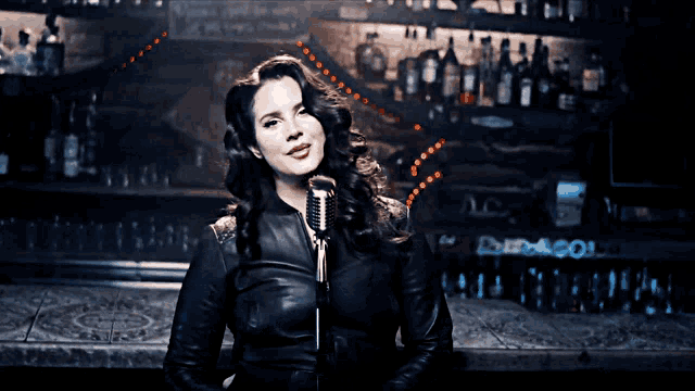a woman in a leather jacket is singing into a microphone in front of a bar
