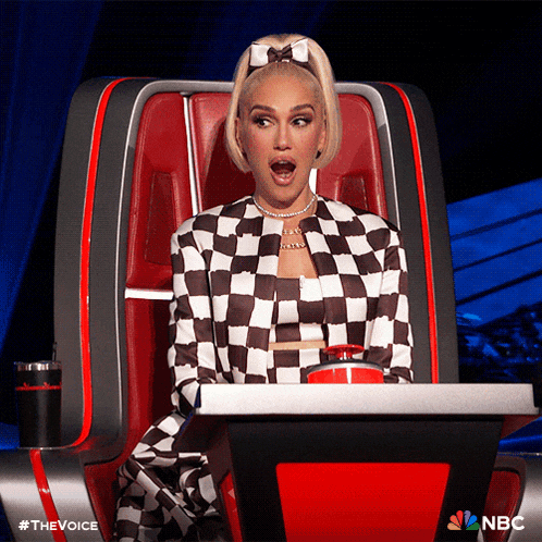 a woman in a checkered outfit is sitting in a chair with a nbc logo behind her