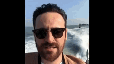 a man with a beard wearing sunglasses is standing on a boat in the water .