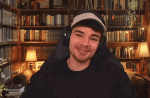 a man wearing headphones and a hat smiles in front of a bookshelf