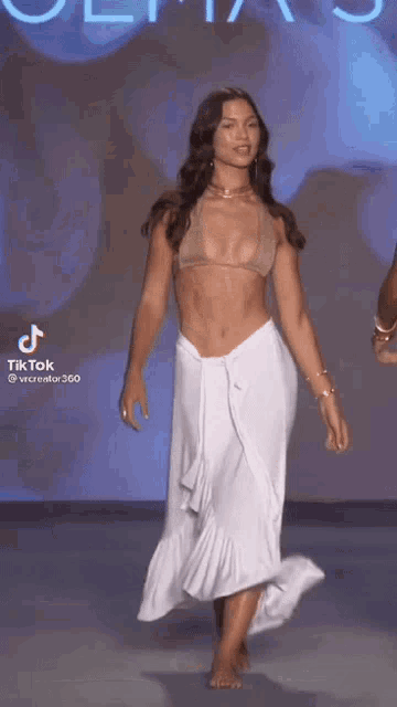a woman is walking down a runway wearing a bikini top and a white skirt .