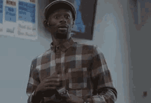a man in a plaid shirt and hat is standing in front of a wall holding a cell phone .