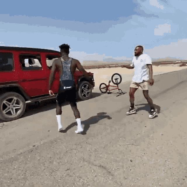 two men are standing in front of a red jeep and a bicycle on the ground .