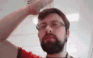 a man with a beard and glasses is standing in front of a white wall and looking at the camera .