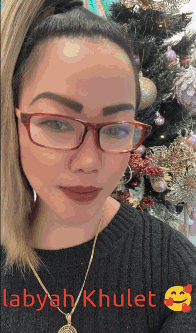 a woman wearing glasses is standing in front of a christmas tree and the name labyah khulet is on the bottom right