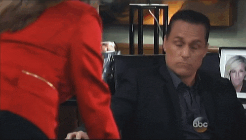 a man in a suit is sitting at a desk with a woman in a red shirt behind him .