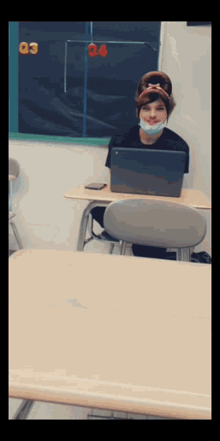 a girl wearing a face mask sits at a desk with a laptop in front of her