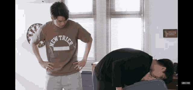 a man wearing a new york t-shirt is standing next to a man laying on a bed .
