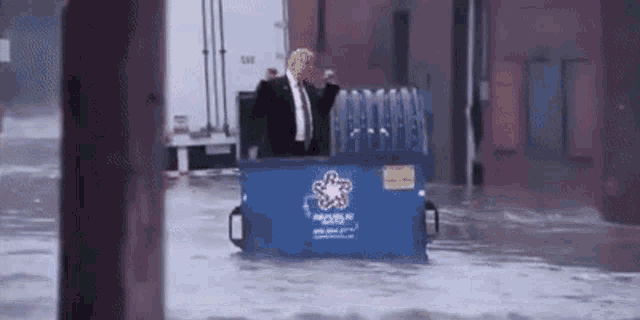 a man in a suit and tie is riding in a dumpster in a flooded street .