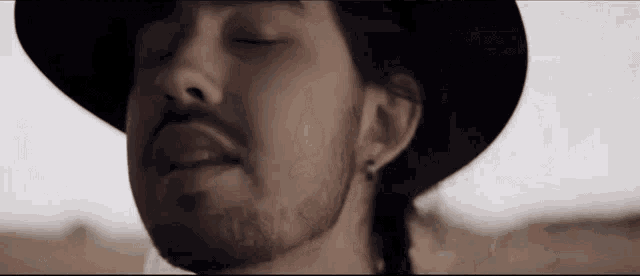 a man with a beard wearing a black hat and earrings is standing in the desert with his eyes closed .