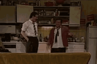 two men are standing next to each other in a kitchen . one of the men is wearing a tie .