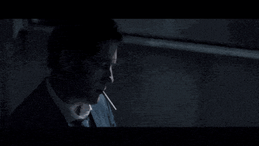 a man in a suit and tie is smoking a cigarette in a dark room