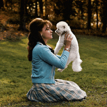 a woman in a blue sweater is kneeling down holding a small white dog