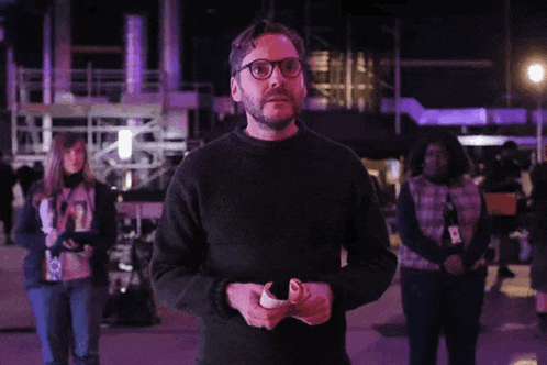 a man with glasses and a beard is standing in front of a crowd holding a piece of paper .