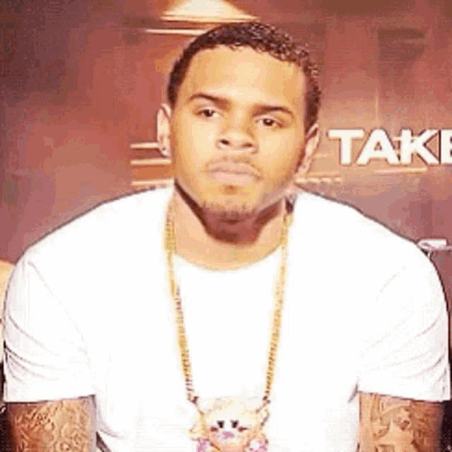 a man wearing a white shirt and a gold chain necklace is sitting in front of a sign that says take .