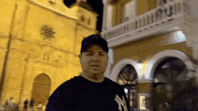 a man wearing a ny yankees shirt is standing in front of a building at night
