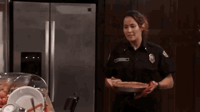 a woman in a firefighter uniform is holding a pie in front of a refrigerator in a kitchen .