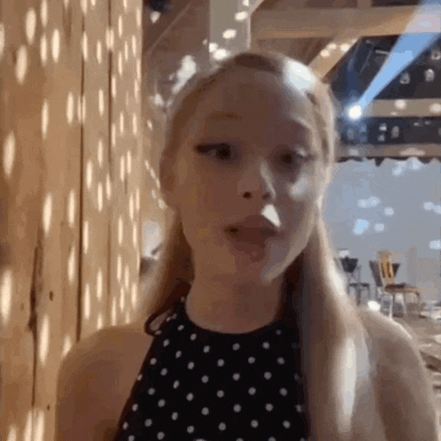 a woman in a black and white polka dot dress is standing in front of a wooden wall and looking at the camera .
