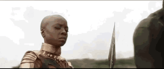 a woman in armor is holding a spear and looking at a man in a field .