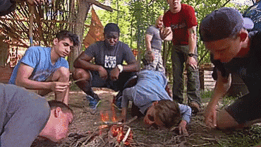a group of young men are gathered around a campfire . one of the men is wearing a shirt that says ikea