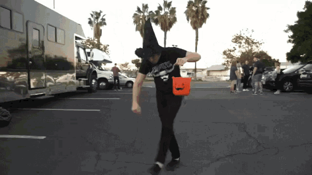 a man in a witch hat holds a trick or treat bucket in a parking lot