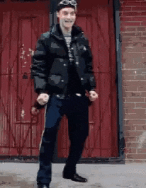 a man in a black jacket and goggles is dancing in front of a red brick building .
