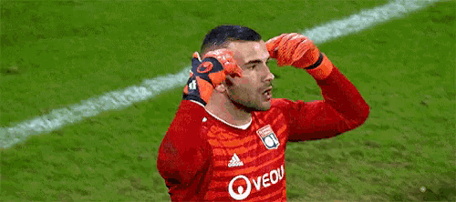 a soccer goalie wearing orange gloves is waving his hand in the air on a field .