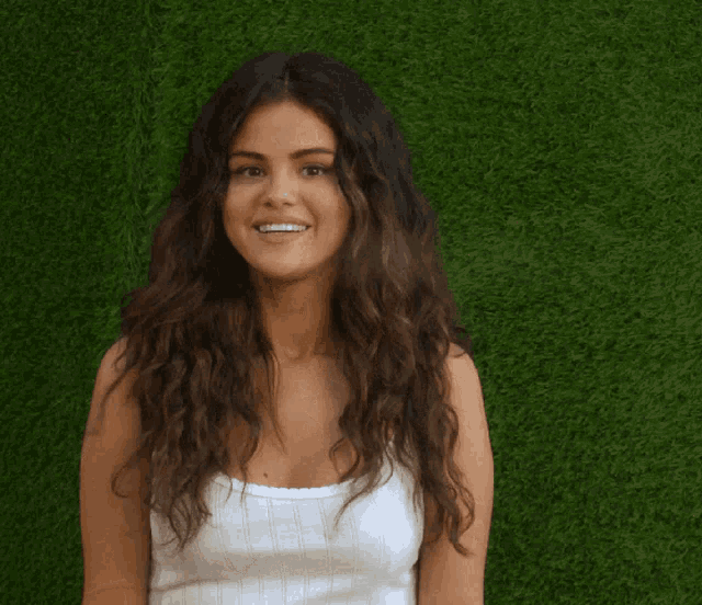 a woman in a white tank top smiles in front of a green background